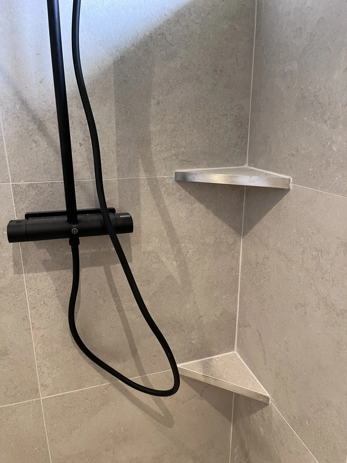 Microcement Halmstad – Duschvägg med fogfri grå yta, svart blandare och hylla i hörnet